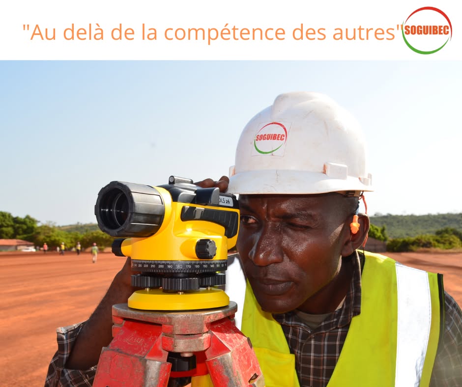 Géomètre-topographe de la SOGUIBEC, casque blanc au logo de l'entreprise, derrière son niveau optique sur un chantier
