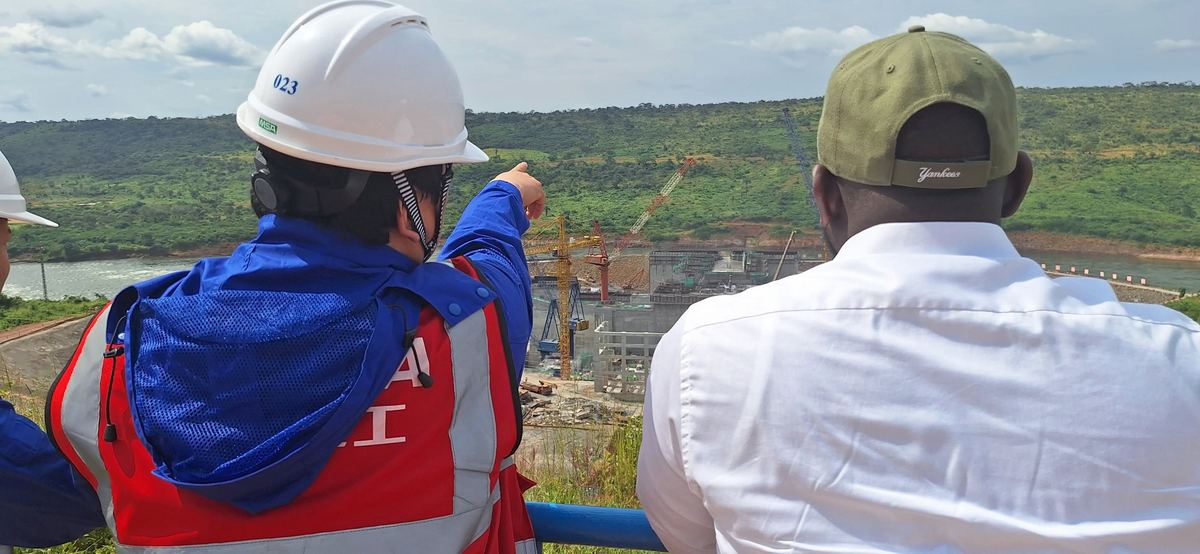 Un ingénieur partenaire désigne un barrage hydroélectrique en construction — grues, ouvrages en béton et fleuve — le Président-Directeur Général à ses côtés