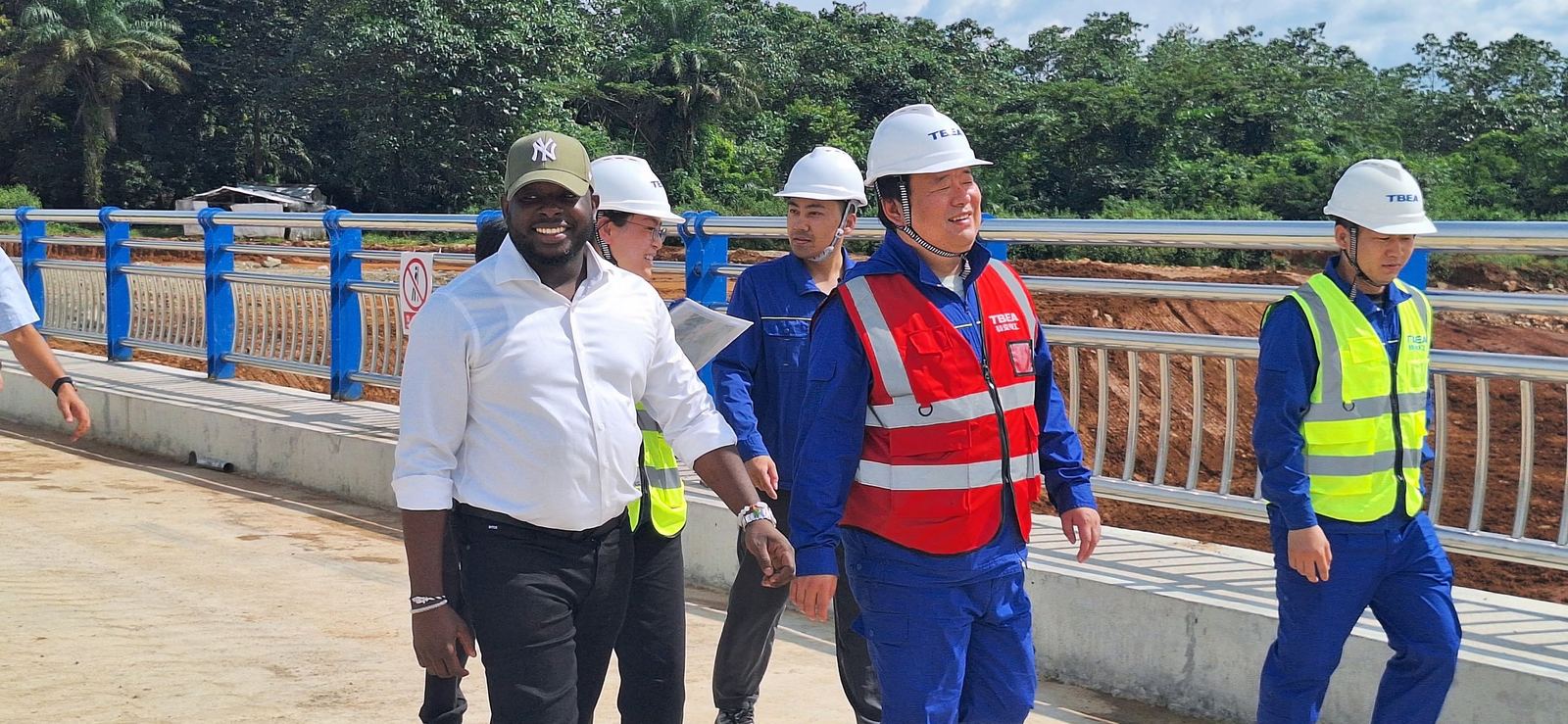 Visite de chantier : le Président-Directeur Général, souriant, traverse un pont achevé aux garde-corps bleus, entouré des partenaires techniques